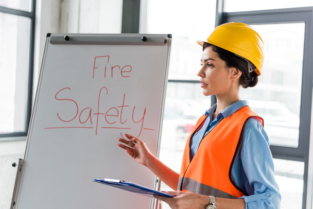  female firefighter in helmet holding clipboard and pen while talking near white board with fire safety lettering - Fotoğraf, Görsel