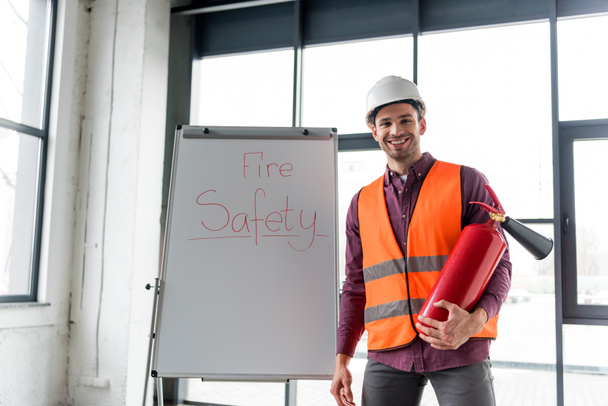 happy fireman holding red extinguisher while standing near white board with fire safety lettering - Photo, Image