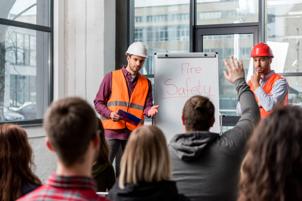 selective focus of handsome firemen in helmets giving talk on briefing near white board with fire safety lettering and looking at man raising hand - Photo, Image
