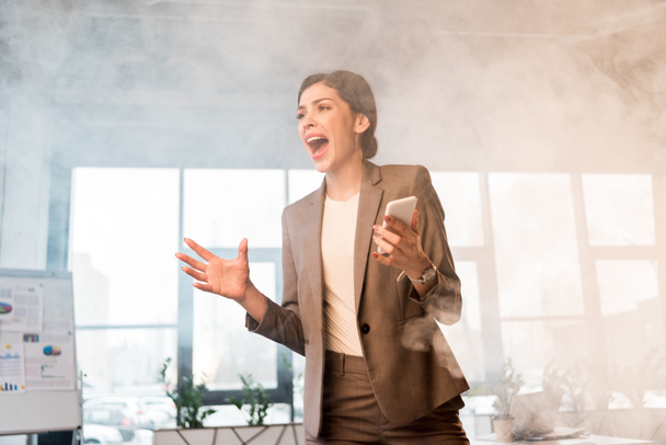 terrified businesswoman screaming whle holding smartphone in office with smoke - Фото, изображение