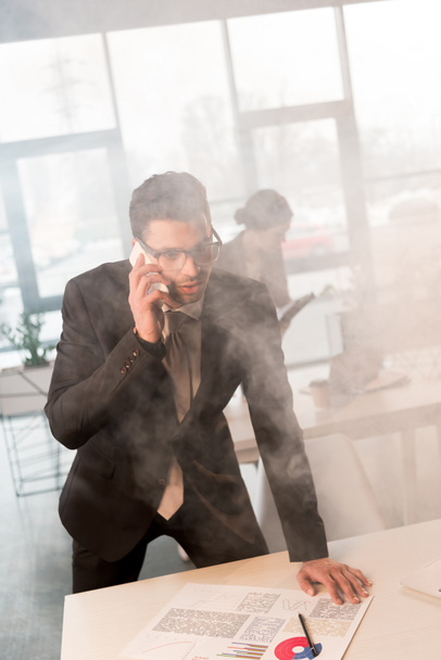 scared businessman in glasses talking on smartphone in office with smoke near coworker - Photo, Image