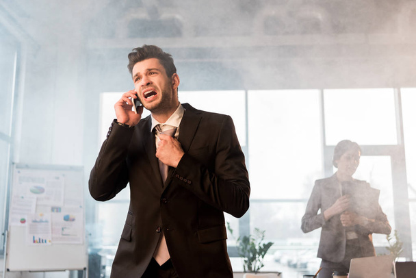 terrified businessman talking on smartphone in office with smoke near coworker - Photo, Image