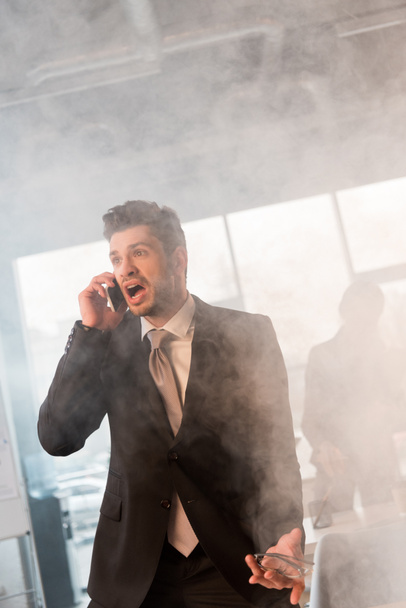 terrified businessman talking on smartphone and screaming in office with smoke near coworker - Foto, afbeelding