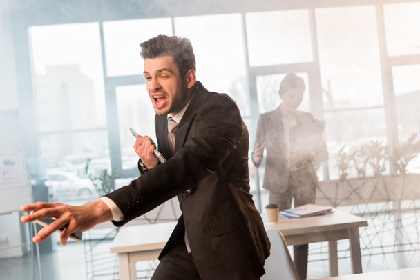 terrified businessman holding smartphone and screaming in office with smoke near female coworker - Photo, image