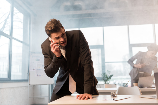 hombre de negocios asustado hablando en el teléfono inteligente en la oficina con humo cerca de compañera de trabajo
 - Foto, Imagen