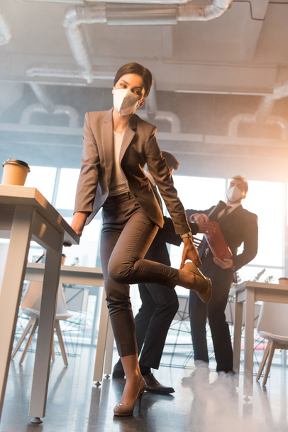 mujer de negocios con máscara que sostiene el zapato mientras está de pie cerca de compañeros de trabajo en la oficina con humo
 - Foto, imagen