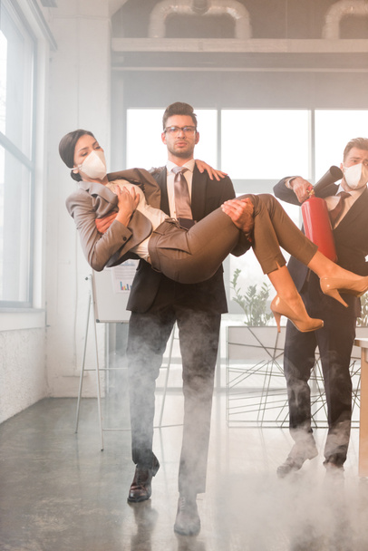 guapo hombre de negocios en gafas sosteniendo en brazos a compañera de trabajo con los ojos cerrados cerca de colega sosteniendo extintor en la oficina con humo
 - Foto, Imagen