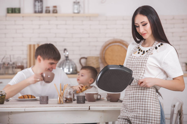 若い母親は、台所に彼女の家族の前に立っています。幸せな家族が夕食や朝食を持っていること。女性が彼女の夫と小さな赤ちゃんのために夕食を作って.  - 写真・画像