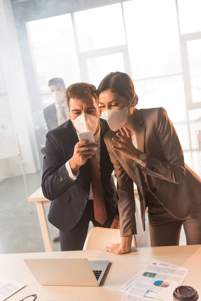 selective focus of terrified colleagues in masks looking at smartphone in office with smoke - Fotografie, Obrázek