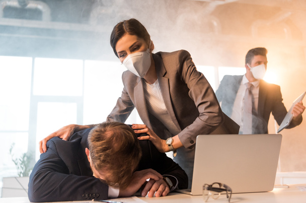 selective focus of terrified woman in mask standing near colleague lying on desk near laptop in office with smoke - Фото, зображення
