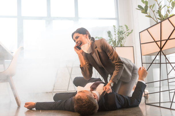 terrified businesswoman in mask talking on smartphone near colleague lying on floor in office with smoke - Foto, afbeelding