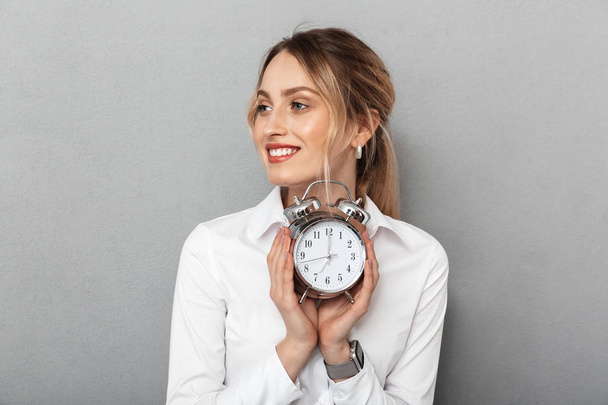 Happy business woman isolated over grey wall background holding alarm clock. - Foto, afbeelding