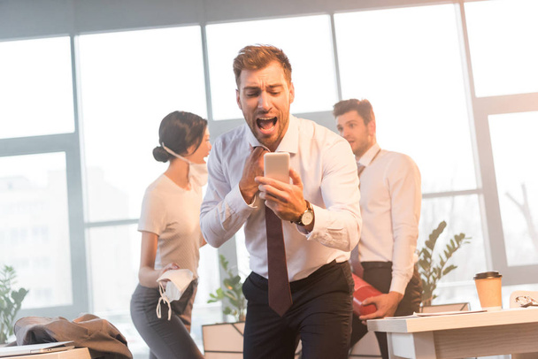 избирательный фокус испуганного бизнесмена, кричащего, глядя на смартфон с коллегами в офисе с дымом на заднем плане
  - Фото, изображение