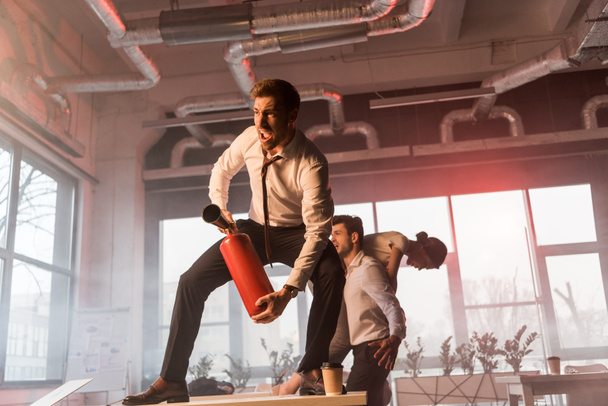 scared businessman yelling while holding extinguisher and standing on desk near coworkers in office with smoke - Photo, Image