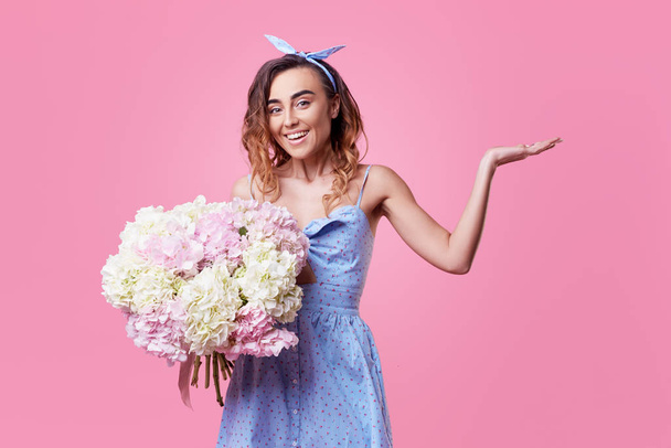 Cheerful young lady in retro round glasses being excited to get bouquet of blue spring flowers on women's day isolated over pink background. It is looking to the right, space for text - 写真・画像