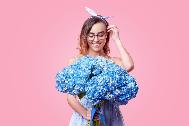 Cheerful young lady in retro round glasses being excited to get bouquet of blue spring flowers on women's day isolated over pink background. It is looking to the right, space for text - Valokuva, kuva