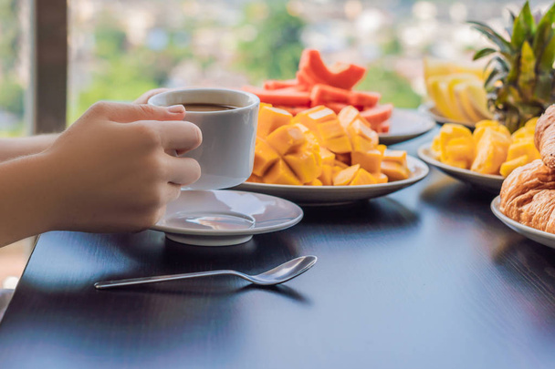 A young woman is having breakfast on the balcony. Breakfast table with coffee fruit and bread croisant on a balcony against the backdrop of the big city - Foto, Imagen