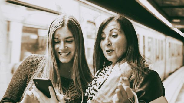 Pari naispuolista ystävää katselemassa puhelinta metroasemalla.
 - Valokuva, kuva