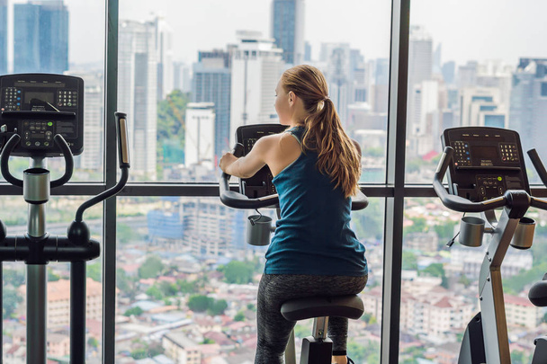 Young woman on a stationary bike in a gym on a big city background - Foto, immagini