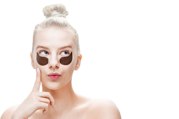 Beautiful young blond woman with black eye patches with a pensive facial expression on a white background isolated, with copy space to insert the product. Care and face masks - Foto, Imagem