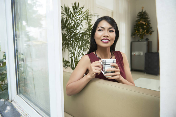 Mujer asiática sonriente disfrutando de su tiempo libre en el sofá con una taza de café y soñando mientras mira por la ventana abierta
 - Foto, Imagen