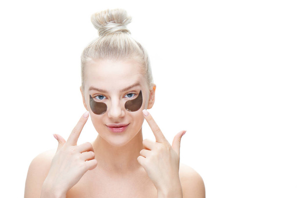Beautiful young blond woman shows fingers on eye patches of black color on a white background in isolation. Care and face masks - Photo, image