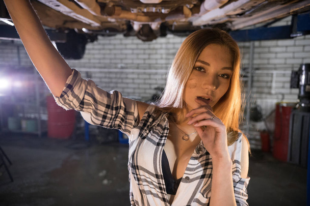 Jonge sexy mechanic vrouw in een unbuttoned shirt reputatie onder de auto in auto reparatieservice. Kijken in de camera - Foto, afbeelding