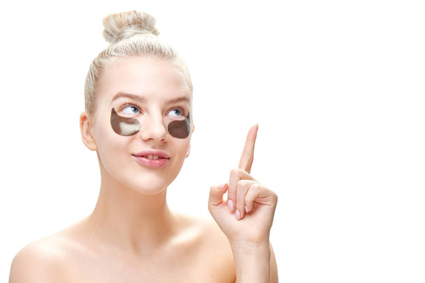 Beautiful young blond woman with blue eyes and full lips and an expression of joy, with black eye patches points the finger at copy space for your product, isolated on white background in isolation. Masks and face care - Photo, image