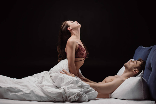attractive young sexy woman in lingerie sitting on top of handsome man in bed isolated on black - Photo, image