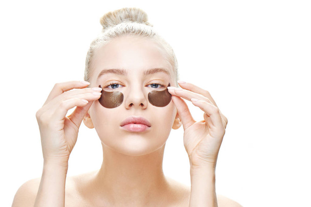 Beautiful young blonde with blue eyes is putting on or removing eye patches on a white background close-up isolated. Masks and face care - 写真・画像