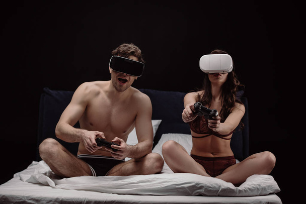 young sexy couple in virtual reality headsets playing video game in bed isolated on black  - Photo, Image