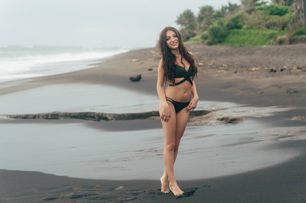 Sexy brunette girl in swimwear resting on black sand beach. Attractive model with gorgeous body posing near ocean - Zdjęcie, obraz