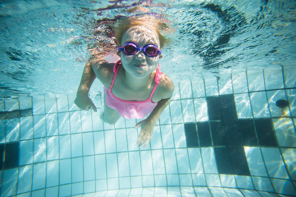 Wide angle underwater photo of a toddler girl swimming in a big swimming pool with goggles and a pink bikini - Photo, Image