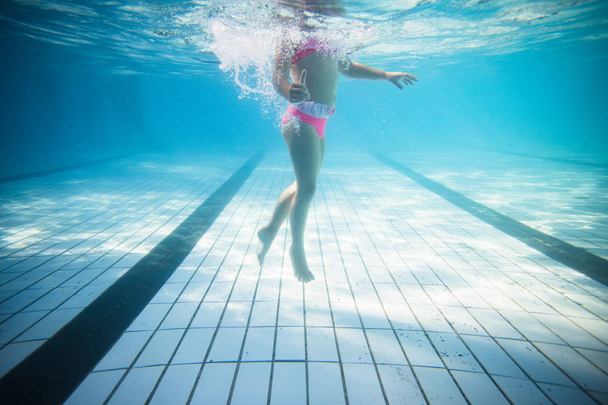 Wide angle underwater photo of a toddler girl swimming in a big swimming pool with goggles and a pink bikini - Foto, Bild