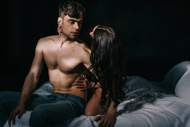 woman embracing handsome shirtless man in bed isolated on black with copy space - Foto, imagen