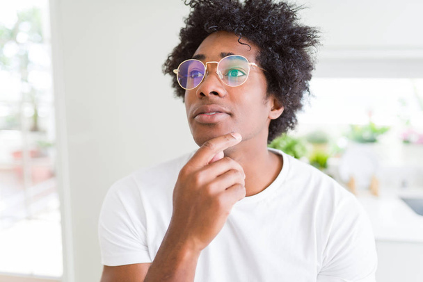 Homem afro-americano usando óculos com a mão no queixo pensando em questão, expressão pensativa. Sorrindo com cara atenciosa. Conceito de dúvida
. - Foto, Imagem