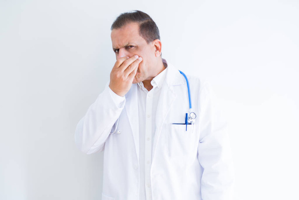 Médecin d'âge moyen homme portant stéthoscope et manteau médical sur fond blanc sentant quelque chose puant et dégoûtant, odeur intolérable, retenant souffle avec les doigts sur le nez. Concept de mauvaises odeurs
. - Photo, image