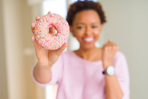 Jovem afro-americana comendo donut de açúcar rosa gritando orgulhoso e celebrando vitória e sucesso muito animado, torcendo emoção
 - Foto, Imagem
