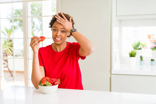 Jonge african american vrouw eten verse aardbeien bij het ontbijt benadrukt met de hand op het hoofd, geschokt met schaamte en verrassing worden geconfronteerd, boos en gefrustreerd. Angst en boos voor fout. - Foto, afbeelding