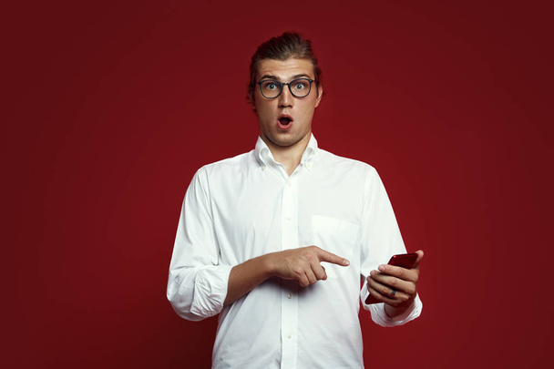 Ritratto di un giovane sorpreso in camicia bianca e occhiali che punta il dito verso il cellulare isolato su sfondo rosso
 - Foto, immagini