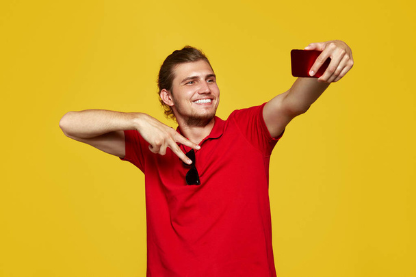 Bild eines glücklichen jungen kaukasischen Mannes, der isoliert auf gelbem Hintergrund posiert, macht ein Selfie per Telefon mit Friedensgeste. - Foto, Bild