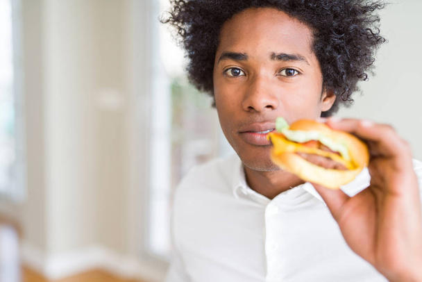 Africká americká hladový člověk jíst hamburger na oběd s výrazem věří na inteligentní tvář myslí vážně - Fotografie, Obrázek