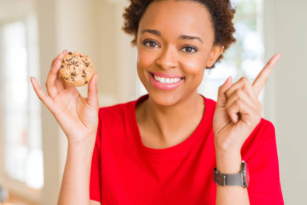 Young african american woman eating chocolate chips cookies very happy pointing with hand and finger to the side - Photo, Image