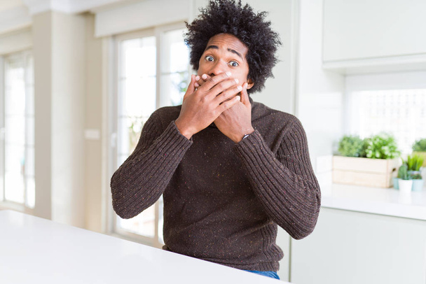 Uomo afroamericano che indossa maglione invernale scioccato copertura bocca con le mani per errore. Concetto segreto
. - Foto, immagini