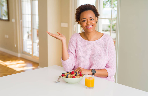 junge afrikanisch-amerikanische Frau, die morgens zu Hause gesund frühstückt, fröhlich lächelnd präsentiert und mit der Hand in die Kamera zeigt. - Foto, Bild