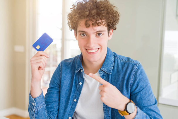 Νέος άνθρωπος που κρατά πολύ χαρούμενος, δείχνοντας με το χέρι και το δάχτυλό της πιστωτικής κάρτας - Φωτογραφία, εικόνα