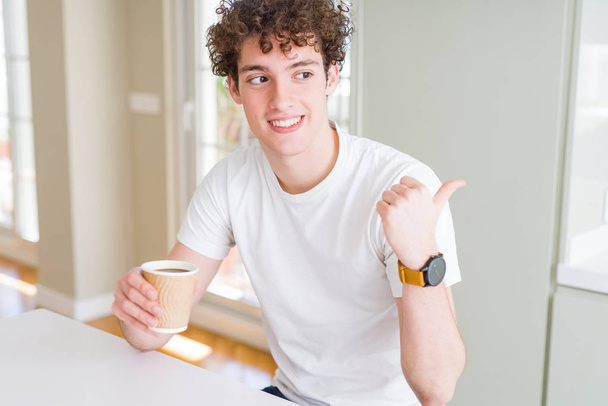 Młody człowiek picia filiżankę kawy w domu wskazując i Wyświetlono z kciukiem do boku z szczęśliwy twarz uśmiechający się zabrać - Zdjęcie, obraz