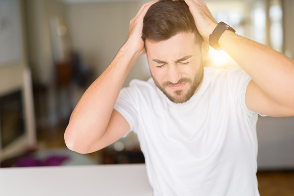 Jonge knappe man dragen casual wit t-shirt in huis lijden aan hoofdpijn wanhopig en benadrukt omdat pijn en migraine. Handen op het hoofd. - Foto, afbeelding