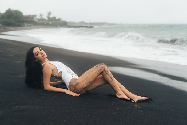 Beautiful sexy girl with long hair lying on beach with black sand. Young woman enjoys relaxing near ocean - Фото, изображение