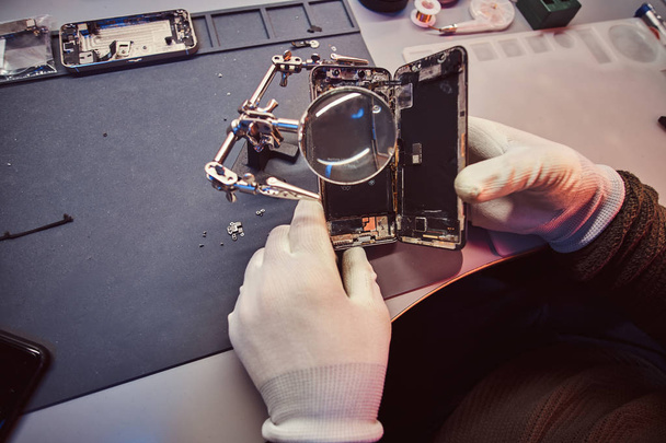 Technician uses a magnifying glass to carefully inspect the internal parts of the smartphone in a modern repair shop - Photo, Image